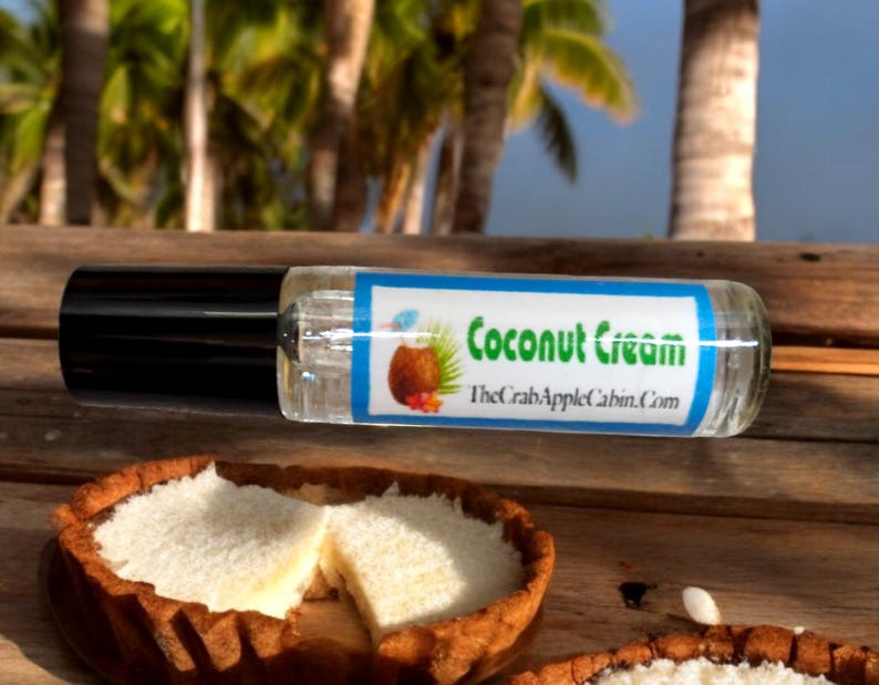 May include: A clear glass roll-on bottle with a black cap and a blue label that says "Coconut Cream" and "TheCrabAppleCabin.com". The bottle is sitting on a wooden surface next to two coconut cream pies.