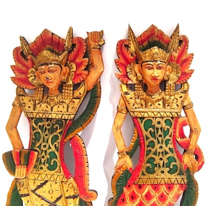 Puede incluir: Dos figuras de madera talladas a mano de bailarines balineses. Cada figura está pintada con detalles dorados, rojos, verdes y naranjas. Las figuras tienen elaborados tocados y posan en posiciones de baile tradicionales. El fondo es blanco.