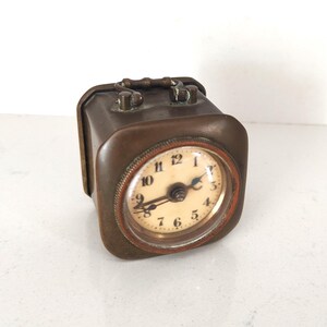 K&ouml;nnte beinhalten: Eine rechteckige Vintage-Reiseuhr mit einem bronzefarbenen Metallgeh&auml;use. Das Zifferblatt ist rund mit schwarzen Ziffern auf cremefarbenem Hintergrund. Sie hat einen Griff oben und einen kleinen, rotbraunen Ring um das Zifferblatt.