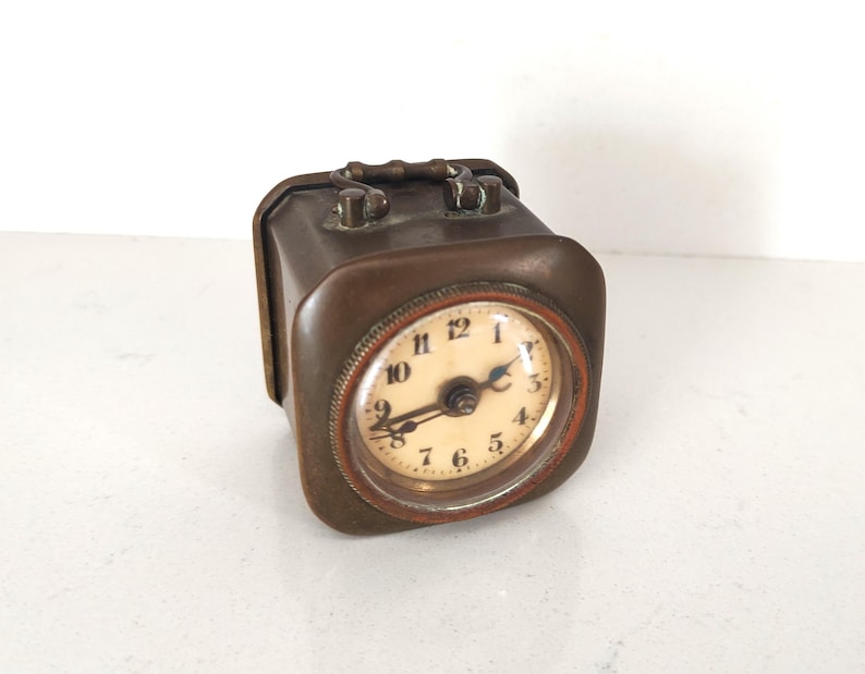 K&ouml;nnte beinhalten: Eine rechteckige Vintage-Reiseuhr mit einem bronzefarbenen Metallgeh&auml;use. Das Zifferblatt ist rund mit schwarzen Ziffern auf cremefarbenem Hintergrund. Sie hat einen Griff oben und einen kleinen, rotbraunen Ring um das Zifferblatt.