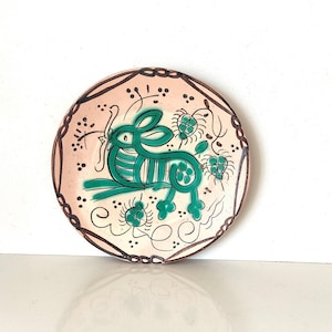 Könnte beinhalten: Eine runde, dekorative Keramikplatte mit handgemaltem Design. Die Platte hat einen hellrosa Hintergrund mit einem braunen Rand. In der Mitte befindet sich ein grüner Hase, umgeben von grünen floralen Akzenten und schwarzen Linien.