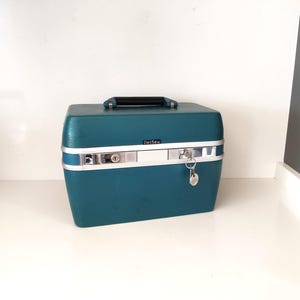 May include: Teal vintage travel case with a black handle and silver accents. The case has a rectangular shape with rounded corners, a silver lock, and a key. The brand name "DelSey" is visible on the front.