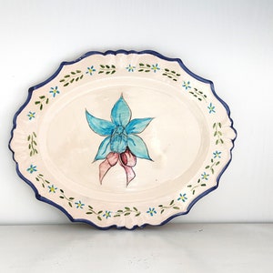 Op de afbeelding: Een witte ovale keramische serveerschaal met een blauw en roze bloemenontwerp. De schaal heeft een gegolfde rand en een blauwe en groene bloemenrand.