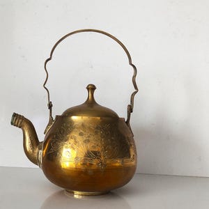 Könnte beinhalten: Eine goldfarbene Metallteekanne mit einem verzierten Design und einem gebogenen Griff. Die Teekanne hat einen Ausguss und einen Deckel.