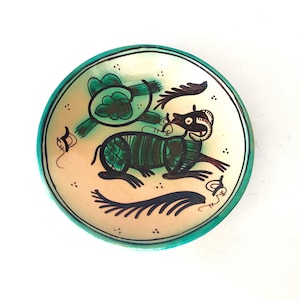 Op de afbeelding: Een ronde keramische kom met een crèmekleurig interieur en een turquoise rand. De kom heeft een handgeschilderd ontwerp van een wezen met een lang lichaam en een schildpadachtige figuur in groen en zwart.