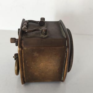 K&ouml;nnte beinhalten: Eine Vintage-Uhr in Messingoptik mit rechteckigem Geh&auml;use und rundem Zifferblatt. Die Uhr hat einen Griff oben und einen kleinen Knopf an der Seite. Das Design deutet auf eine klassische, antike &Auml;sthetik hin.