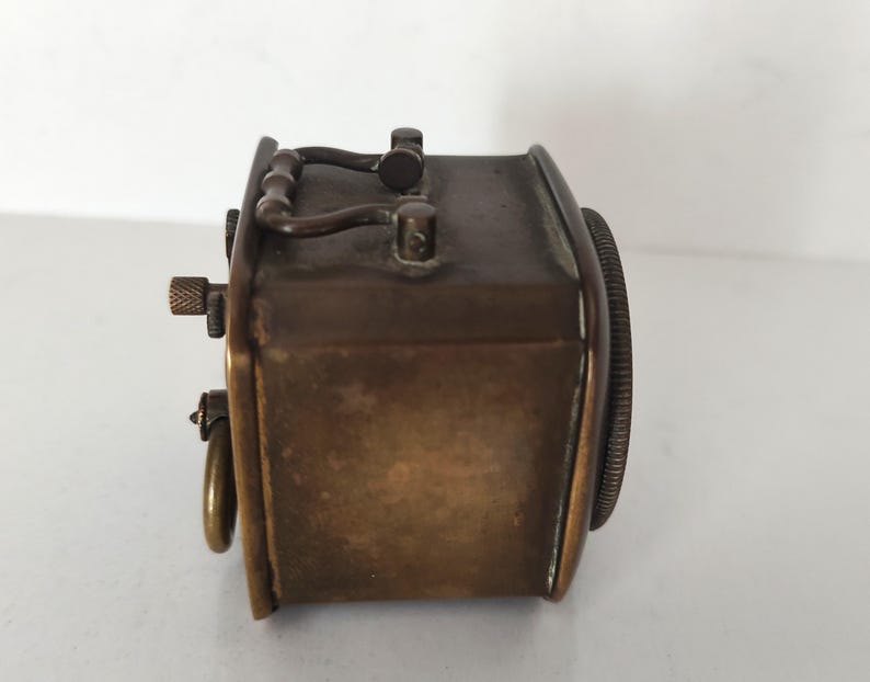 K&ouml;nnte beinhalten: Eine Vintage-Uhr in Messingoptik mit rechteckigem Geh&auml;use und rundem Zifferblatt. Die Uhr hat einen Griff oben und einen kleinen Knopf an der Seite. Das Design deutet auf eine klassische, antike &Auml;sthetik hin.