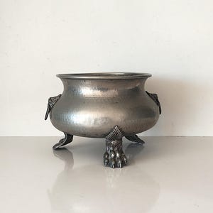 May include: A hammered metal bowl with three claw feet and decorative handles. The bowl has a wide, rounded shape and a silver finish. The handles are ornate, and the feet are detailed. The bowl is likely used for decorative purposes.