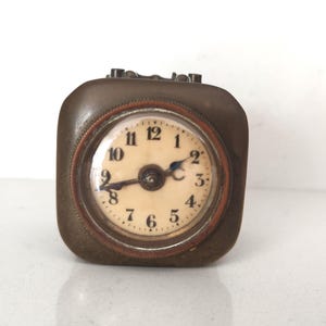 K&ouml;nnte beinhalten: Eine quadratische Vintage-Reiseuhr mit einem bronzefarbenen Metallgeh&auml;use. Das Zifferblatt ist cremefarben mit schwarzen Ziffern und Zeigern. Sie hat ein kleines, rundes Zifferblatt mit Glasabdeckung und braunem Rand.