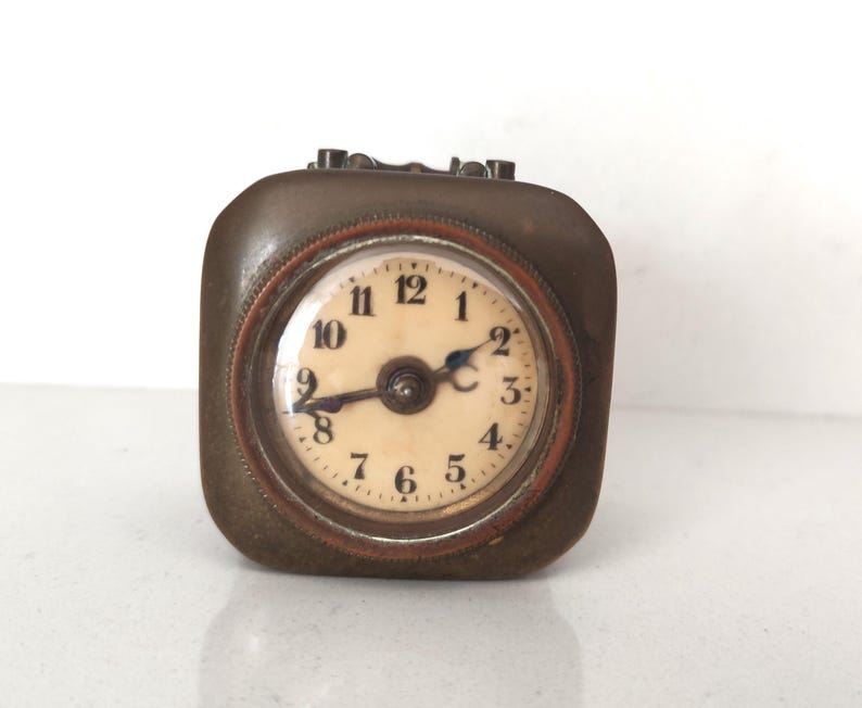 K&ouml;nnte beinhalten: Eine quadratische Vintage-Reiseuhr mit einem bronzefarbenen Metallgeh&auml;use. Das Zifferblatt ist cremefarben mit schwarzen Ziffern und Zeigern. Sie hat ein kleines, rundes Zifferblatt mit Glasabdeckung und braunem Rand.