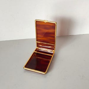 Puede incluir: Una caja de baratijas rectangular abierta con un patrón de carey en el interior y un marco dorado. La caja está hecha de un material brillante y está colocada sobre un fondo neutro.