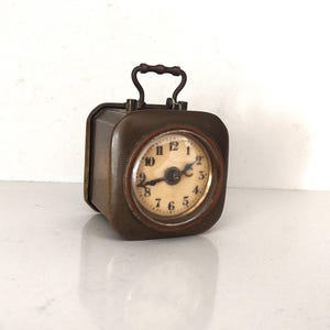 K&ouml;nnte beinhalten: Eine rechteckige Vintage-Reiseuhr mit Griff. Die Uhr hat ein rundes Zifferblatt mit schwarzen Ziffern auf cremefarbenem Hintergrund. Das Metallgeh&auml;use der Uhr hat eine dunkle, gealterte Oberfl&auml;che. Die Uhr ist etwa 7.6 cm hoch.