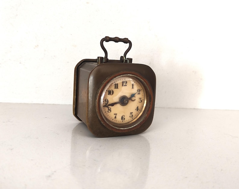 K&ouml;nnte beinhalten: Eine rechteckige Vintage-Reiseuhr mit Griff. Die Uhr hat ein rundes Zifferblatt mit schwarzen Ziffern auf cremefarbenem Hintergrund. Das Metallgeh&auml;use der Uhr hat eine dunkle, gealterte Oberfl&auml;che. Die Uhr ist etwa 7.6 cm hoch.