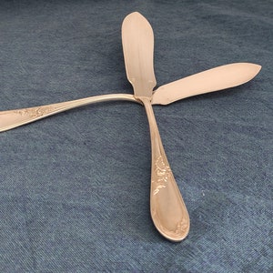 May include: Two silver butter knives with ornate handles, lying on a blue fabric surface.