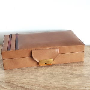 Könnte beinhalten: Eine rechteckige, hellbraune Lederbox mit Klappdeckel und goldfarbenem Verschluss. Die Oberseite der Box weist drei vertikale Streifen in Braun- und Burgundertönen auf. Die Box steht auf einer Holzoberfläche.