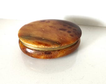 May include: Oval-shaped, lidded trinket box with a warm, mottled brown finish. The lid and base are connected by a thin, gold-colored band. The box has a glossy, polished appearance, suggesting it is made of a hard material.