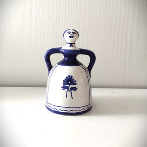 May include: A white ceramic bell shaped like a woman with blue painted details. The woman has a blue dress with a white flower design and a blue face with a smile.