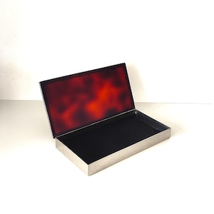 May include: A rectangular metal box with a hinged lid featuring a mottled reddish-brown tortoiseshell pattern. The interior of the box is lined with a solid black fabric, and the exterior frame has a smooth, metallic silver-toned finish.