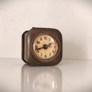 K&ouml;nnte beinhalten: Eine quadratische Vintage-Reiseuhr mit einem bronzefarbenen Metallgeh&auml;use. Das Zifferblatt ist cremefarben mit schwarzen Ziffern und Zeigern. Die Uhr ist etwa 5 cm breit und 5 cm hoch.
