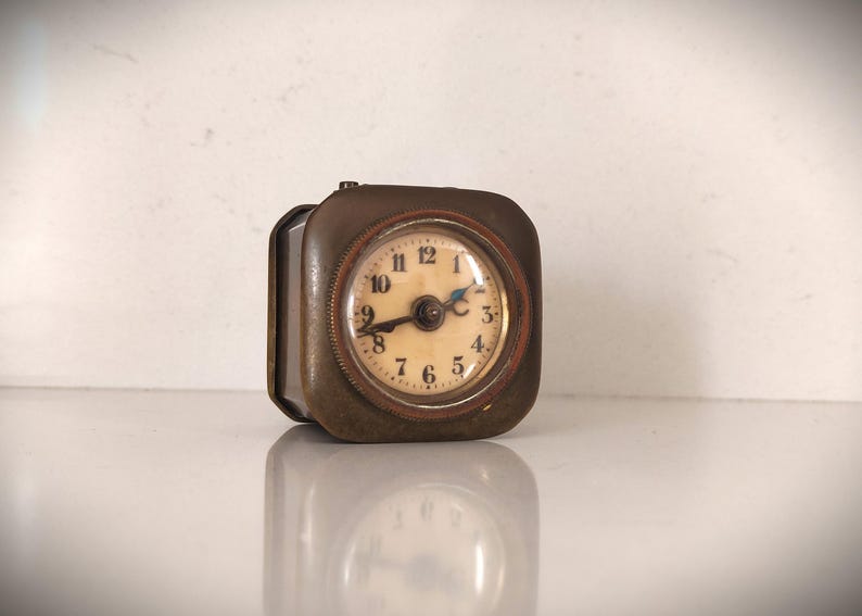 K&ouml;nnte beinhalten: Eine quadratische Vintage-Reiseuhr mit einem bronzefarbenen Metallgeh&auml;use. Das Zifferblatt ist cremefarben mit schwarzen Ziffern und Zeigern. Die Uhr ist etwa 5 cm breit und 5 cm hoch.