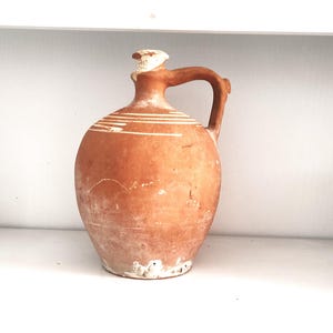 Puede incluir: Una jarra de terracota con un esmalte blanco y un asa. La jarra tiene un cuerpo redondo y un cuello estrecho.