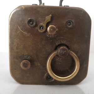 K&ouml;nnte beinhalten: Nahaufnahme der R&uuml;ckseite eines Vintage-Reiseweckers aus Messing. Das Zifferblatt der Uhr zeigt einen Aufziehschl&uuml;ssel, einen kleinen Ring und die W&ouml;rter "REVE" und "HEURE". Die Uhr hat eine rechteckige Form.