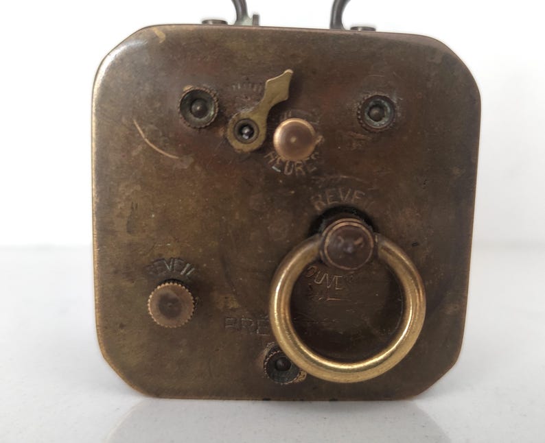 K&ouml;nnte beinhalten: Nahaufnahme der R&uuml;ckseite eines Vintage-Reiseweckers aus Messing. Das Zifferblatt der Uhr zeigt einen Aufziehschl&uuml;ssel, einen kleinen Ring und die W&ouml;rter "REVE" und "HEURE". Die Uhr hat eine rechteckige Form.