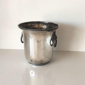 Könnte beinhalten: Silberner Weinkühler mit Seilgriffen. Der Kühler hat eine breite Öffnung und einen dekorativen Rand. Das Metall hat ein leicht angelaufenes Aussehen, was seinen Vintage-Charme unterstreicht. Ein klassisches Stück für gekühlte Getränke.