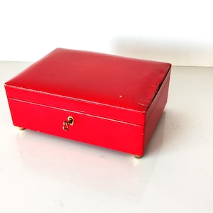 May include: A red rectangular jewellery box with a keyhole and a gold-coloured key. The box is set on small, gold-coloured feet. The box has a slightly worn appearance.