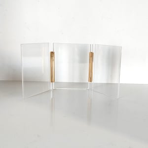 Pode incluir: Um suporte de exibição de acrílico transparente com três painéis, conectados por dobradiças verticais de metal dourado. O suporte foi projetado para dobrar e desdobrar. Os painéis são retangulares e ficam em pé.
