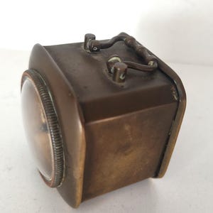 K&ouml;nnte beinhalten: Eine Vintage-Reiseuhr in Messingoptik mit gew&ouml;lbtem Glaszifferblatt und geriffelter L&uuml;nette. Die Uhr hat einen rechteckigen K&ouml;rper mit abgerundeten Ecken und einem Griff. Das Design deutet auf einen klassischen, antiken Stil mit sichtbarem Zifferblatt hin.