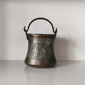 Op de afbeelding: Een antieke metalen emmer met een decoratieve handgreep. De emmer heeft een reliëfontwerp rond de body en een geschulpte rand. Het metaal heeft een aangetaste, verouderde uitstraling, met tinten brons en zilver.