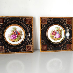 以下が含まれることがあります： 金色の縁取りと装飾的なデザインが施された正方形の黒いフレームが2つ。それぞれのフレームには、庭園の風景の中に描かれたカップルが描かれた白い磁器のプレートが入っています。