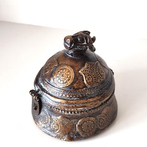Puede incluir: Una caja de baratijas decorativa de color bronce con intrincados patrones en relieve y un pequeño asa ornamentada. La caja presenta una tapa con un diseño detallado y una pequeña figura decorativa en la parte superior. Estética vintage.