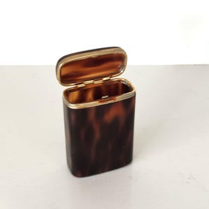 May include: A rectangular case with a hinged lid featuring a brown and black tortoiseshell pattern. The container has a metallic gold-toned rim and hinge mechanism, with a smooth, polished finish on the exterior and interior surfaces.