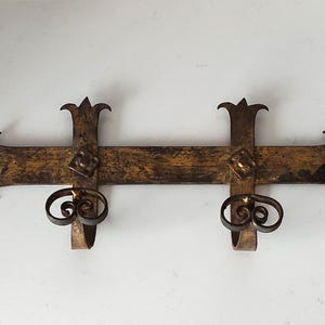 Könnte beinhalten: Ein antiker Wandleuchter aus Metall mit zwei Kerzenhaltern. Der Leuchter hat eine dunkle, gealterte Goldoberfläche mit dekorativen Schnörkeln und spitzen Zierknospen. Das Design ist symmetrisch und hat einen horizontalen Balken mit vertikalen Stützen.
