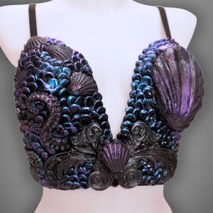 Könnte beinhalten: Ein bauchfreies Oberteil mit tiefem V-Ausschnitt, verziert mit irisierenden, schuppenartigen Applikationen in Blau und Violett, dunklen, strukturierten Schnörkeln und verschiedenen Muscheln. Mit dünnen schwarzen Trägern.