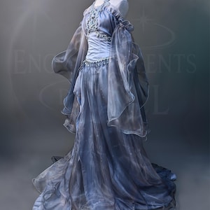 May include: A long, flowing gown in blue and grey tones, shown on a mannequin. The dress has a halter neck, detailed bodice, and sheer, layered sleeves. The skirt falls to the floor, creating a dramatic look. Suitable for fantasy or formal occasions, the dress is elegant and ethereal.
