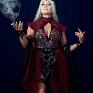Peut inclure: Une robe rouge foncé et noire dotée d'un col montant, d'ornements en filigrane argenté sur le corsage et de panneaux en tissu transparent superposés. La tenue comprend une coiffe noire et de longues manches fluides avec des bracelets.