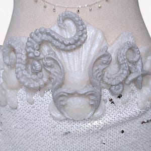Könnte beinhalten: Ein weißes paillettenbesetztes Oberteil mit einem Muschel- und Oktopus-Tentakel-Design. Über dem Oberteil hängt eine silberne Kette mit kleinen, funkelnden Verzierungen. Das Design ist aufwendig und detailliert, mit einem perlmuttartigen Finish.
