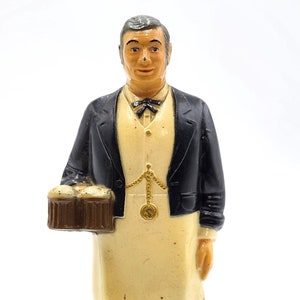 May include: A vintage figurine of a man in a black suit and apron holding a barrel of beer. The base of the figurine has the text "Schmidt's Beer of Philadelphia Ale".