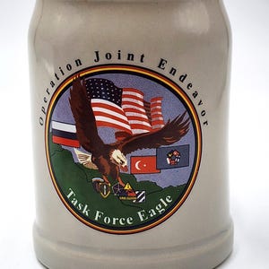 May include: A light gray ceramic beer stein featuring a circular graphic with the text Operation Joint Endeavor and Task Force Eagle. The design includes a bald eagle, the American flag, and several military unit insignias over a green map.