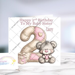 Może przedstawiać: Kwadratowa kartka okolicznościowa z napisem Happy 2nd Birthday To My Baby Sister Lacey. Projekt zawiera dużą cyfrę dwa z przeszywanym wzorem patchworkowym w kolorze beżowym i r&oacute;żowym, małe pudełko prezentowe oraz puszystego szarego misia z r&oacute;żowym sercem.