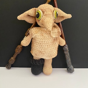 Może przedstawiać: Maskotka Dobby'ego wykonana na szydełku z serii Harry Potter, z dużymi zielonymi oczami, brązowymi włosami z włóczki i czarnymi butami na szydełku. Maskotka stoi obok dwóch drewnianych różdżek.
