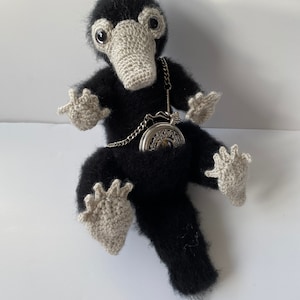 May include: A black and gray crocheted stuffed animal with a long tail and a pocket watch hanging from its neck. The animal has large, round eyes and a long, pointed beak.