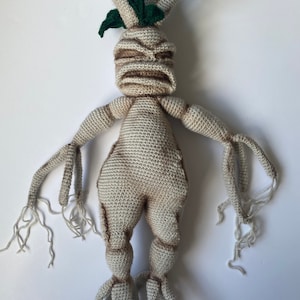 May include: A crocheted plush toy of a humanoid tree creature with a grumpy expression. The toy has a beige body with brown details, green leaves, and long, thin roots.