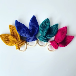 May include: Four crocheted bunny rabbit teethers with wooden rings. The teethers are in yellow, blue, green, and pink.