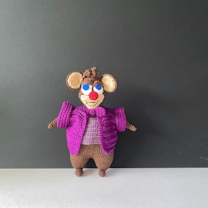 May include: A crocheted brown mouse wearing a purple cardigan and a purple bow tie. The mouse has blue eyes and a red nose.