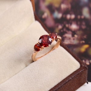 May include: A rose gold ring featuring three red gemstones. The ring has a simple band and the gemstones are set in a row.