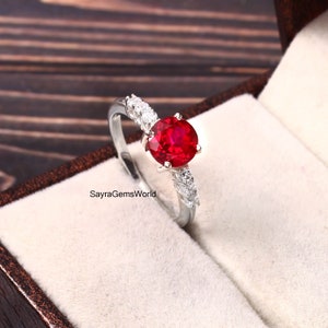 Peut inclure: Une bague en argent avec une grande pierre de rubis rouge ronde entourée de petites pierres claires. La bague est dans un écrin à bague en velours blanc.